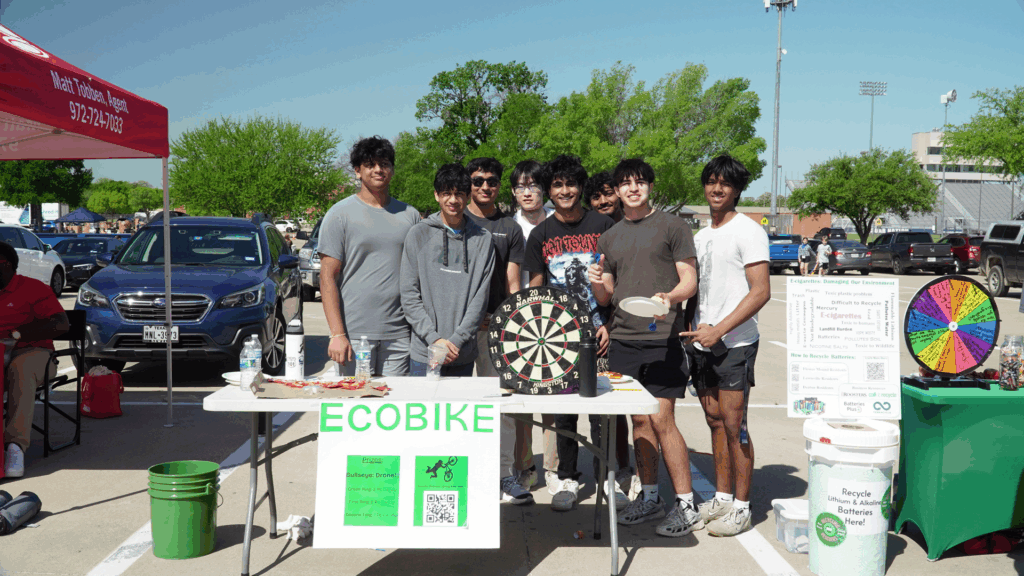 Eight young men of various ethnicities stand behind a table with a sign on it that says ECOBIKE. On the table is a dart board. The table is in a parking lot with other tables with activities on them on each side.