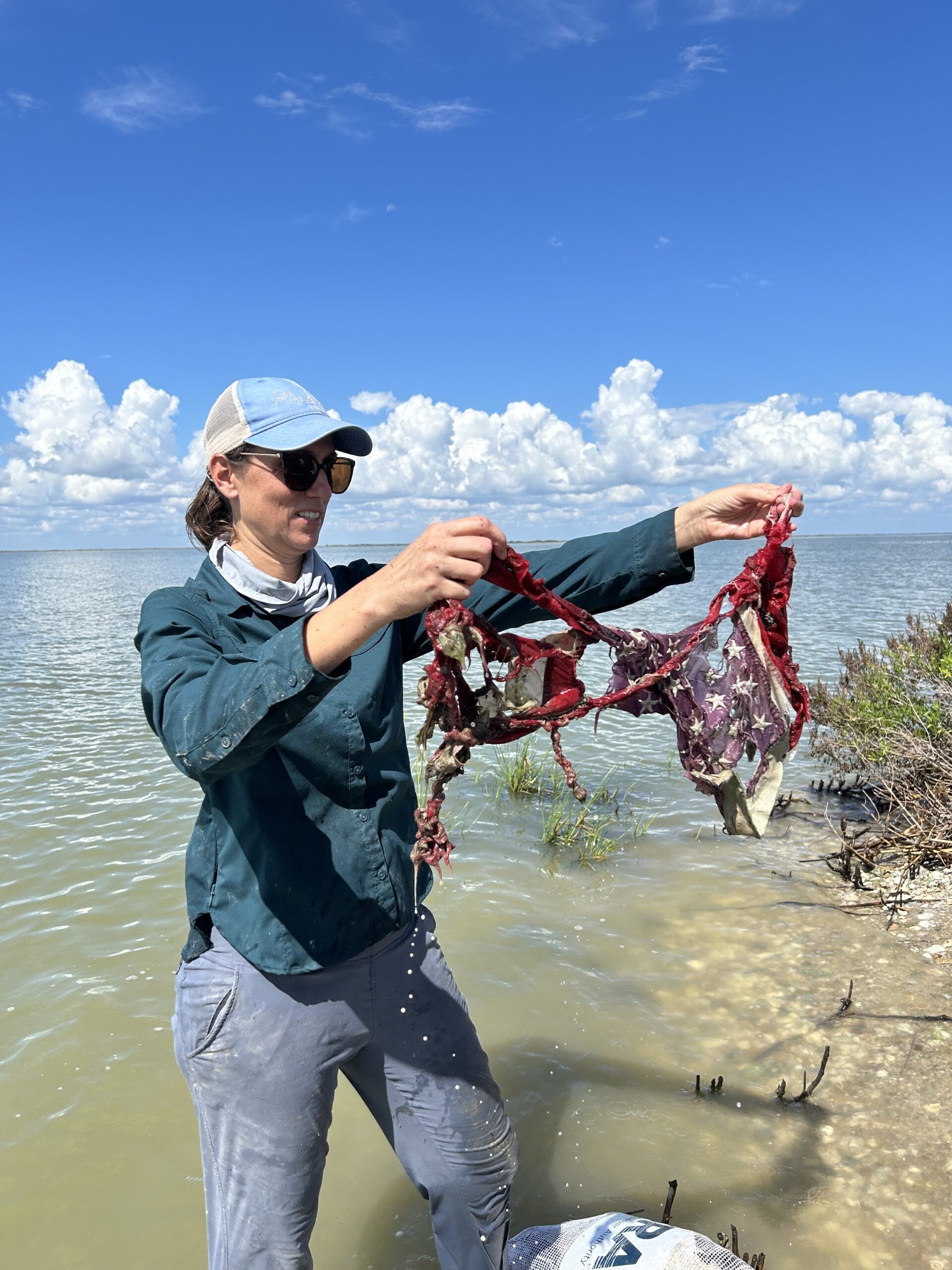 A white woman in sunglasses and a ballcap stands in the water near the shore, holding up a ravaged US flag